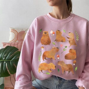 May include: A pink sweatshirt with a graphic print of capybaras in various poses. The capybaras are brown and have small ears and short tails. The print also includes mushrooms and leaves.