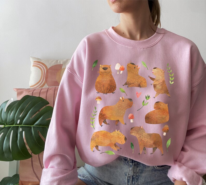 May include: A pink sweatshirt with a graphic print of capybaras in various poses. The capybaras are brown and have small ears and short tails. The print also includes mushrooms and leaves.