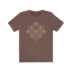 May include: A brown t-shirt with a geometric design featuring a butterfly and crescent moons in a beige color.