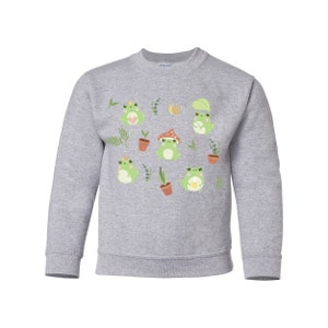 K&ouml;nnte beinhalten: Ein graues Sweatshirt mit einem Muster aus gr&uuml;nen Fr&ouml;schen, Pflanzen und Pilzen. Die Fr&ouml;sche tragen Kronen, H&uuml;te und halten Blumen.