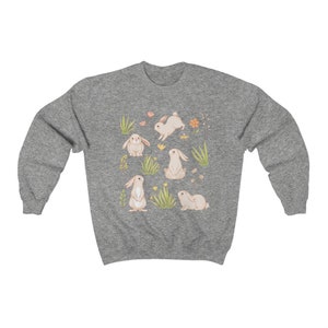 May include: A heather grey sweatshirt with a cute pattern of white bunnies and green grass. The bunnies are in different poses, some are hopping, some are sitting, and some are standing. There are also small yellow flowers scattered throughout the design.