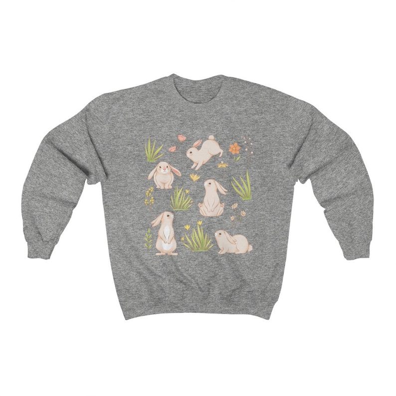 May include: A heather grey sweatshirt with a cute pattern of white bunnies and green grass. The bunnies are in different poses, some are hopping, some are sitting, and some are standing. There are also small yellow flowers scattered throughout the design.