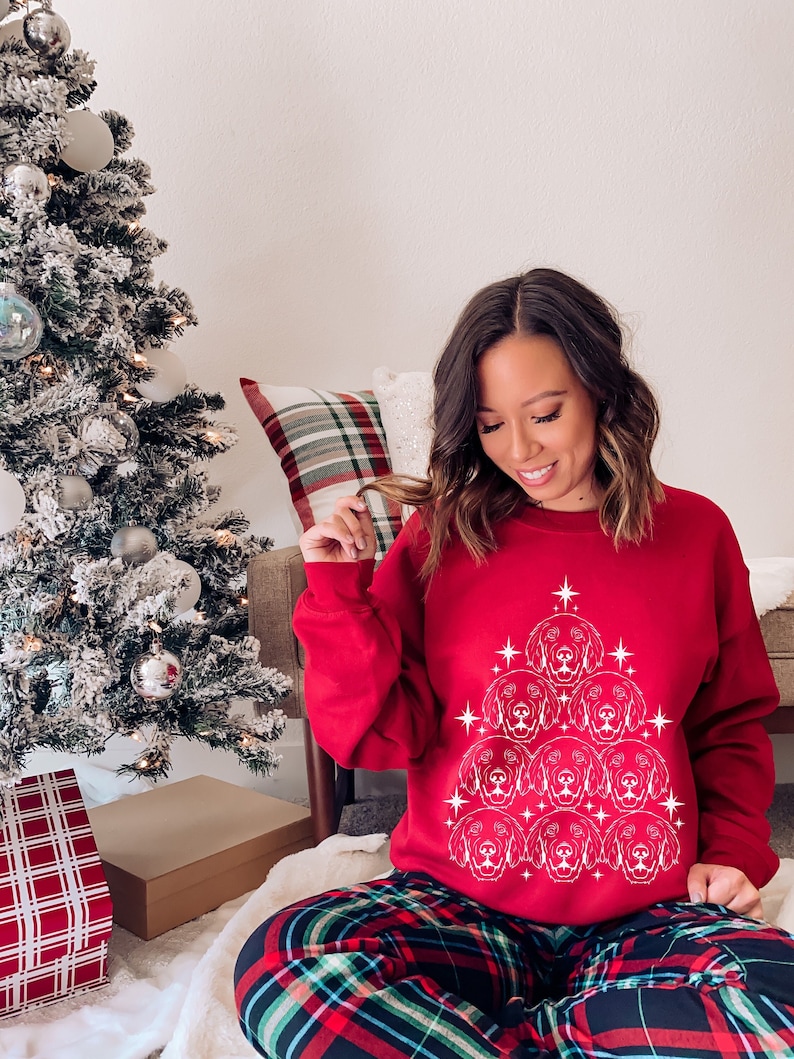 May include: A red Christmas sweater with a white design of a Christmas tree made of dog faces. The sweater is worn by a person sitting on a floor with a plaid pattern.