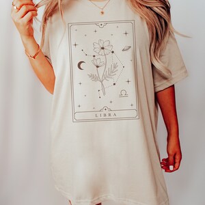 May include: A beige oversized t-shirt with a graphic design of a flower with a Libra zodiac symbol and the word "LIBRA" printed on the front.