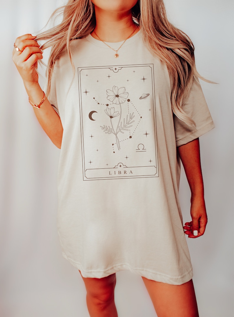 May include: A beige oversized t-shirt with a graphic design of a flower with a Libra zodiac symbol and the word "LIBRA" printed on the front.