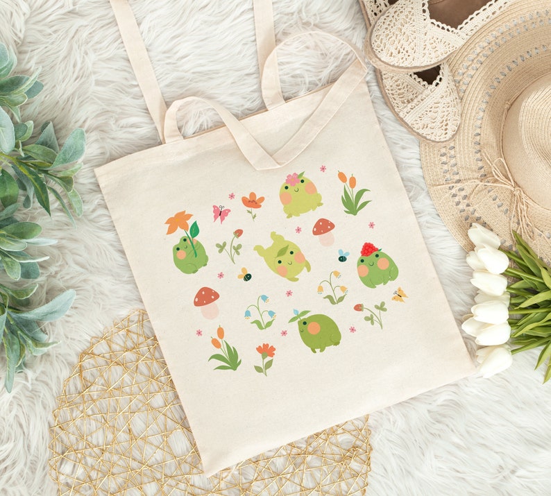 May include: A white canvas tote bag with a design of green frogs, flowers, and mushrooms. The frogs are wearing hats and are surrounded by colorful flowers and mushrooms.