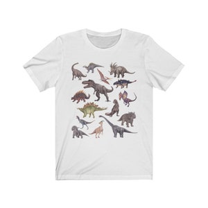 May include: White t-shirt with a colorful illustration of various dinosaurs, including a Tyrannosaurus Rex, Triceratops, Stegosaurus, and Pterodactyl.