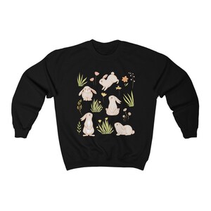 May include: Black sweatshirt with a white bunny rabbit and floral pattern. The bunnies are in different poses, including sitting, standing, and hopping. The flowers are in shades of pink, yellow, and orange.