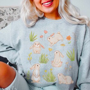 May include: A gray sweatshirt with a graphic print of bunnies and flowers. The bunnies are white with pink noses and ears. The flowers are pink, orange, and yellow. The sweatshirt is a comfortable and stylish choice for spring.