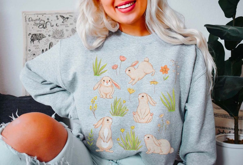 May include: A gray sweatshirt with a graphic print of bunnies and flowers. The bunnies are white with pink noses and ears. The flowers are pink, orange, and yellow. The sweatshirt is a comfortable and stylish choice for spring.