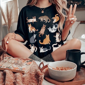 May include: Black t-shirt featuring various cat illustrations in different poses and colors. The shirt is worn by a person sitting on a bed with a tray holding a bowl of cereal and a black mug. A book is also on the bed.