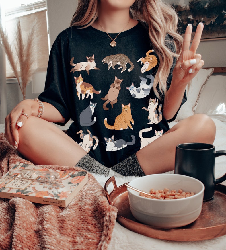 May include: Black t-shirt featuring various cat illustrations in different poses and colors. The shirt is worn by a person sitting on a bed with a tray holding a bowl of cereal and a black mug. A book is also on the bed.