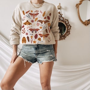 Puede incluir: Una sudadera de color crema con un estampado colorido de polillas, cristales y hojas. La sudadera tiene un corte holgado y un cuello redondo.