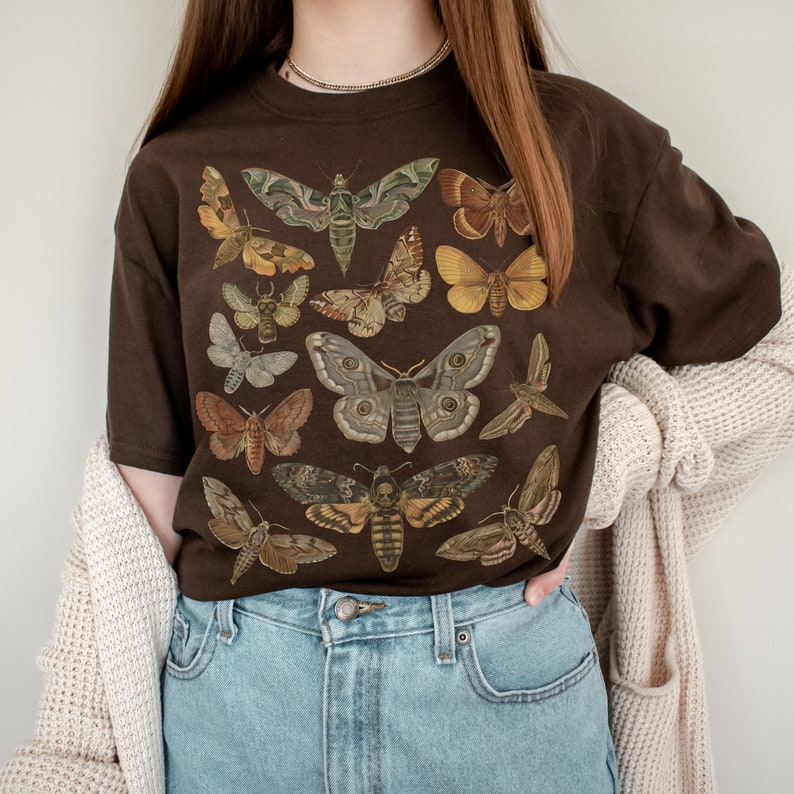 May include: A brown t-shirt with a vintage illustration of various butterflies and moths. The shirt features a variety of colors and patterns, including brown, black, white, yellow, and red.