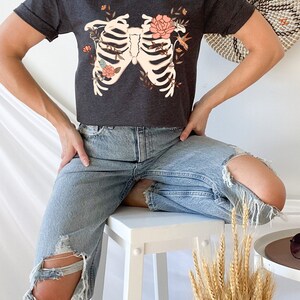 May include: A dark gray t-shirt with a floral ribcage design. The design features a white ribcage with pink flowers and a hummingbird.