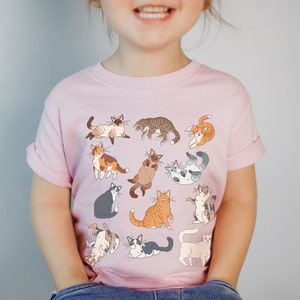 May include: A pink t-shirt with a pattern of various cats in different poses. The cats are drawn in a cartoon style and are in shades of brown, white, gray, and orange.