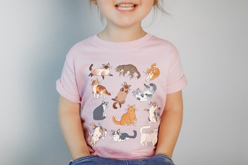 May include: A pink t-shirt with a pattern of various cats in different poses. The cats are drawn in a cartoon style and are in shades of brown, white, gray, and orange.