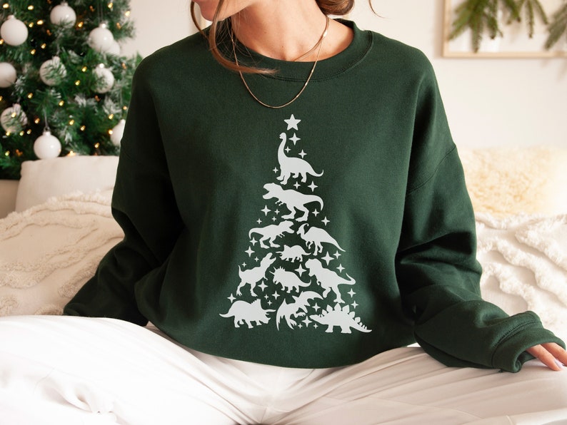 May include: A dark green sweatshirt with a white dinosaur Christmas tree design. The tree is made up of various dinosaurs, including a T-Rex, Triceratops, and Stegosaurus.