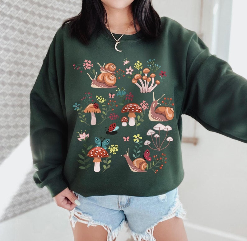 Puede incluir: Una sudadera verde oscuro con un dise&ntilde;o colorido de caracoles, setas y mariposas. El dise&ntilde;o es un patr&oacute;n caprichoso e inspirado en la naturaleza.