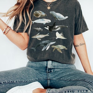 May include: A dark gray t-shirt with a watercolor design of various sea creatures, including a whale, dolphin, sea turtle, and stingray. The text "Mer Oc&eacute;an" is tattooed on the wearer's arm.