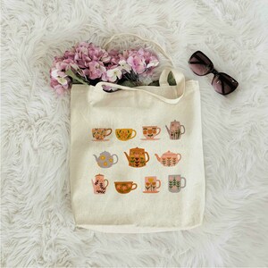 May include: A white canvas tote bag with a floral pattern of teapots and teacups. The design features a variety of colors, including pink, yellow, green, and blue.