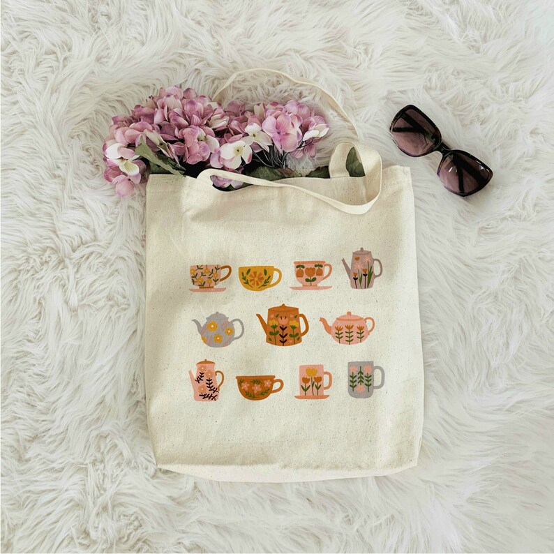 May include: A white canvas tote bag with a floral pattern of teapots and teacups. The design features a variety of colors, including pink, yellow, green, and blue.