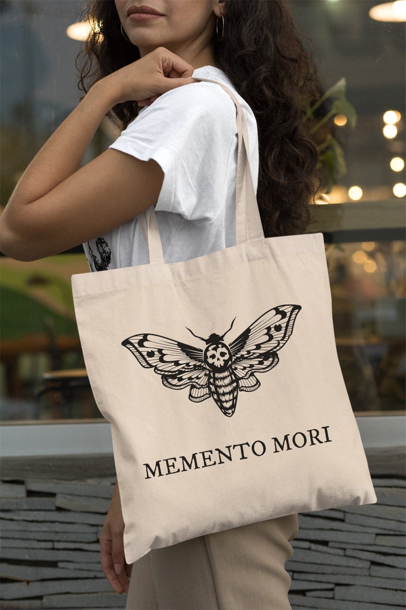 May include: A beige tote bag with black straps features a black and white illustration of a moth with a skull on its body. The text "MEMENTO MORI" is printed in black below the moth.