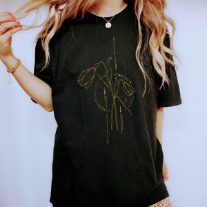 May include: A black oversized t-shirt with a gold floral design. The design features stylized flowers with geometric lines and dots.