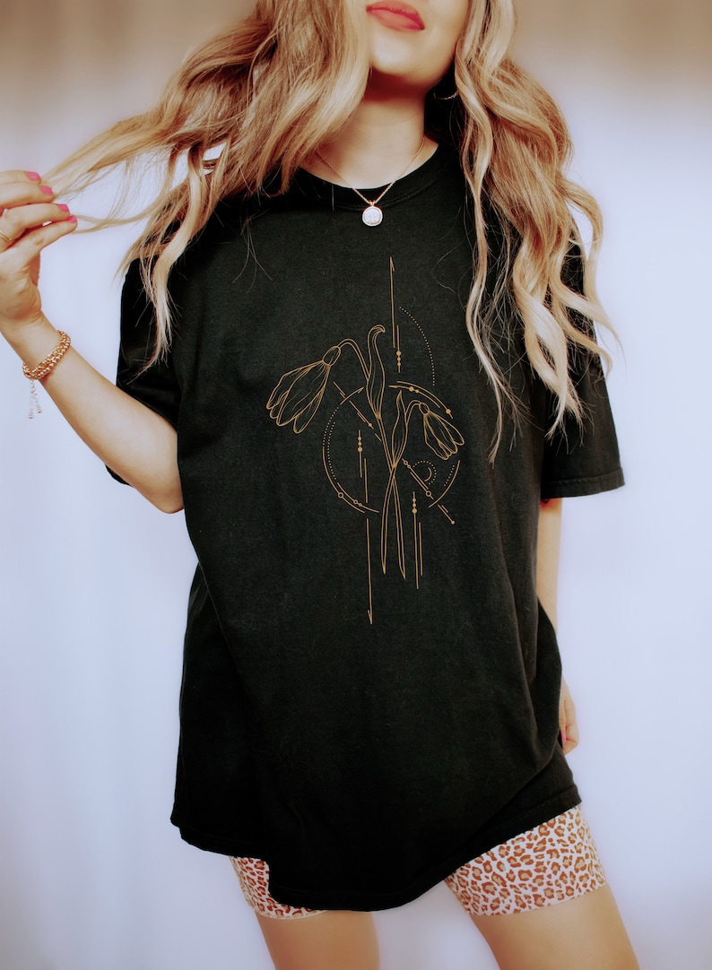 May include: A black oversized t-shirt with a gold floral design. The design features stylized flowers with geometric lines and dots.