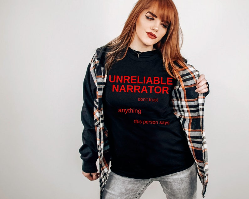 Puede incluir: Una camiseta negra con el texto "UNRELIABLE NARRATOR don't trust anything this person says" en letras rojas. La persona lleva una camisa de cuadros sobre la camiseta.