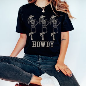K&ouml;nnte beinhalten: Ein schwarzes T-Shirt mit einem Aufdruck von drei Skeletten, die Cowboyh&uuml;te und Stiefel tragen. Der Text "HOWDY" ist unter den Skeletten aufgedruckt.