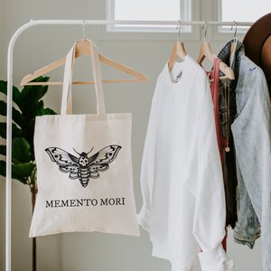 May include: A white canvas tote bag with a black and white moth graphic and the text "MEMENTO MORI". The bag is hanging on a white rack with other clothing items.