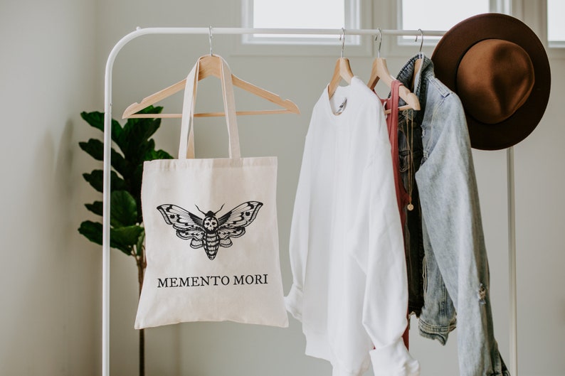 May include: A white canvas tote bag with a black and white moth graphic and the text "MEMENTO MORI". The bag is hanging on a white rack with other clothing items.