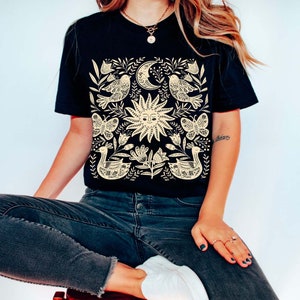 May include: A black t-shirt with a cream-colored design featuring a sun, moon, birds, butterflies, and floral elements. The design is centered on the shirt.