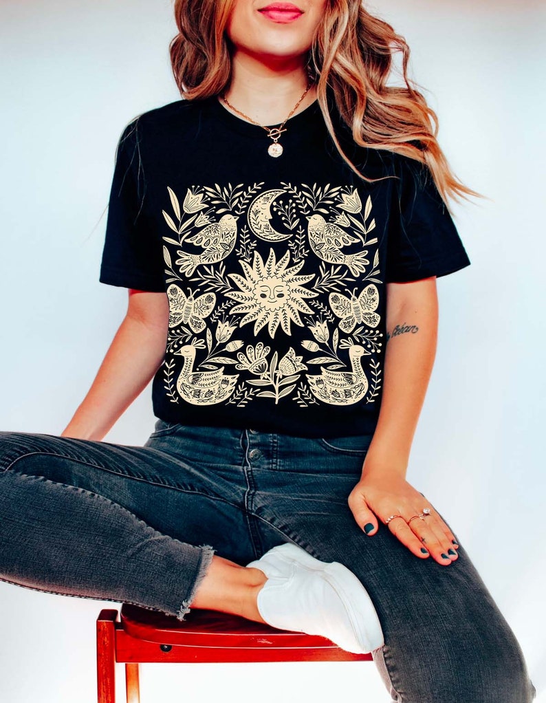 May include: A black t-shirt with a cream-colored design featuring a sun, moon, birds, butterflies, and floral elements. The design is centered on the shirt.