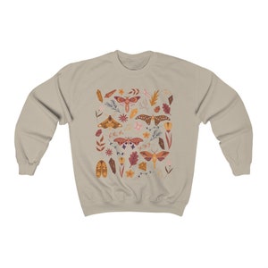 Puede incluir: Una sudadera beige con un patr&oacute;n de polillas, cristales, hojas y flores en tonos marrones, naranjas y rosas.