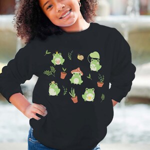 K&ouml;nnte beinhalten: Schwarzes Sweatshirt mit einem gr&uuml;nen Frosch-Design. Die Fr&ouml;sche tragen Kronen, H&uuml;te und halten eine Tasse. Auf dem Sweatshirt sind auch Pflanzen und eine Schnecke zu sehen.