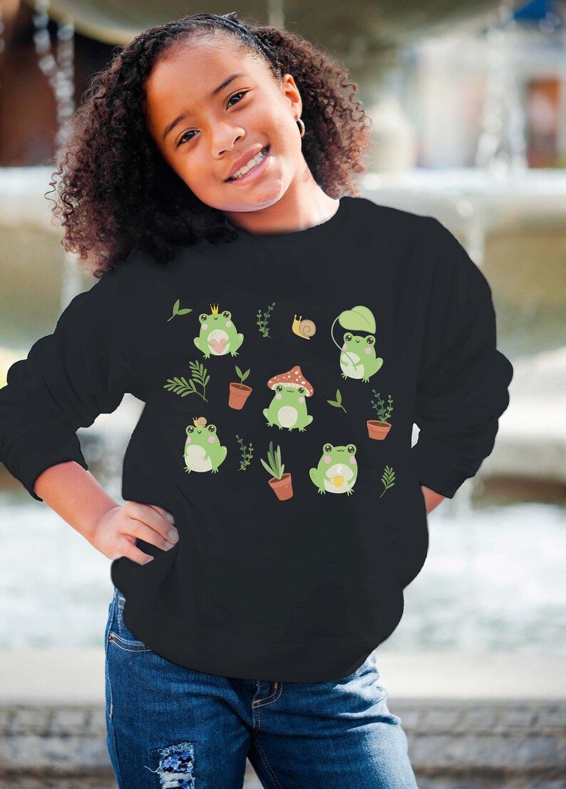 K&ouml;nnte beinhalten: Schwarzes Sweatshirt mit einem gr&uuml;nen Frosch-Design. Die Fr&ouml;sche tragen Kronen, H&uuml;te und halten eine Tasse. Auf dem Sweatshirt sind auch Pflanzen und eine Schnecke zu sehen.