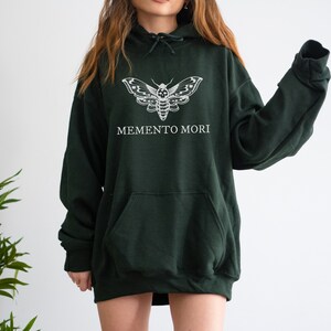 K&ouml;nnte beinhalten: Ein dunkelgr&uuml;ner Kapuzenpullover mit einem wei&szlig;en Motten-Motiv und dem Aufdruck "MEMENTO MORI" auf der Vorderseite.