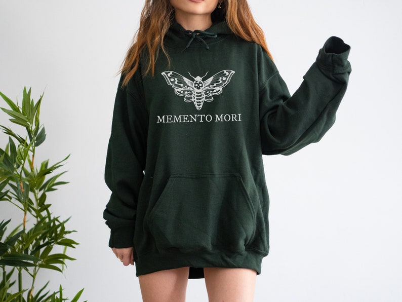 K&ouml;nnte beinhalten: Ein dunkelgr&uuml;ner Kapuzenpullover mit einem wei&szlig;en Motten-Motiv und dem Aufdruck "MEMENTO MORI" auf der Vorderseite.