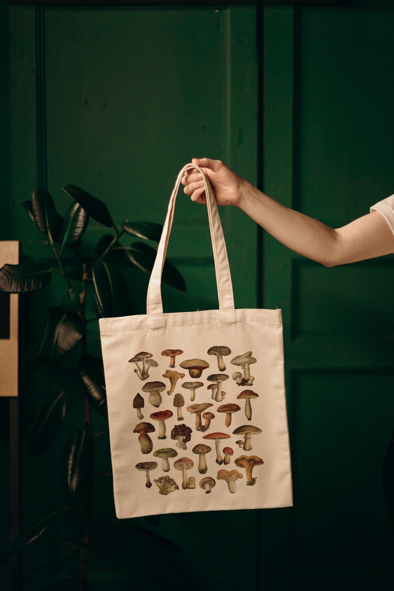 May include: A white canvas tote bag with a design of various mushrooms in different colors and sizes. The bag has two long handles.