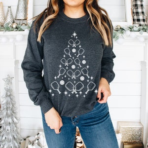 K&ouml;nnte beinhalten: Ein dunkelgraues Sweatshirt mit einem wei&szlig;en Design eines Weihnachtsbaums aus Pickleball-Schl&auml;gern und B&auml;llen.