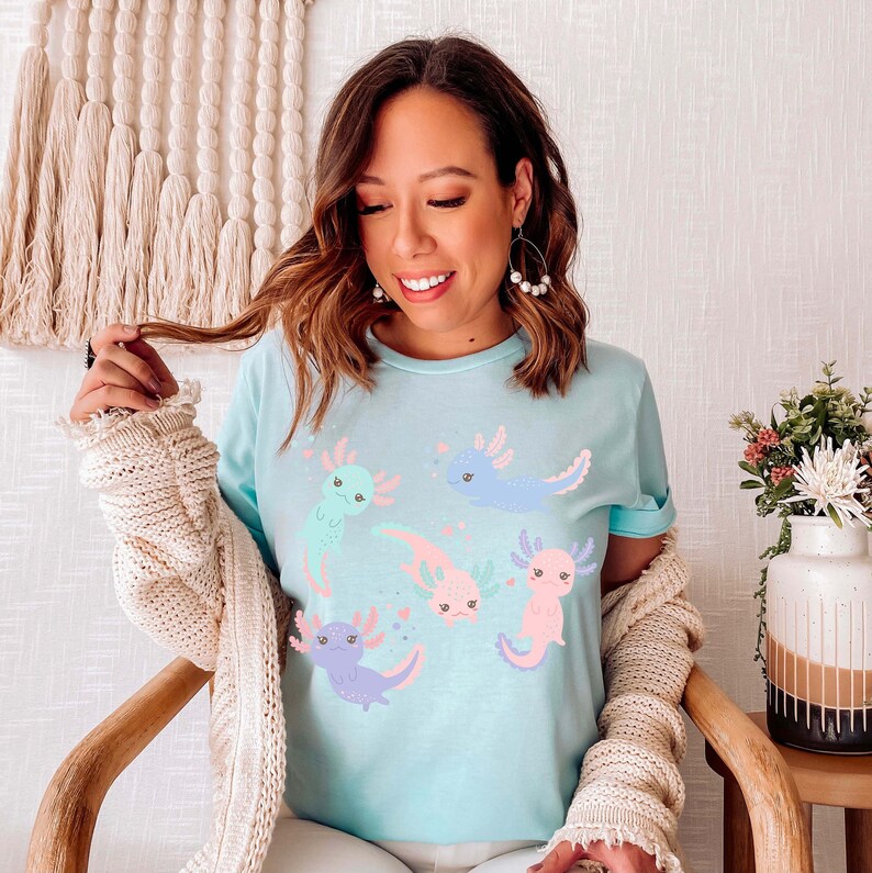 May include: A light blue t-shirt featuring a graphic print of five pink, blue and green axolotls. The axolotls are cartoon-like and have smiling faces.