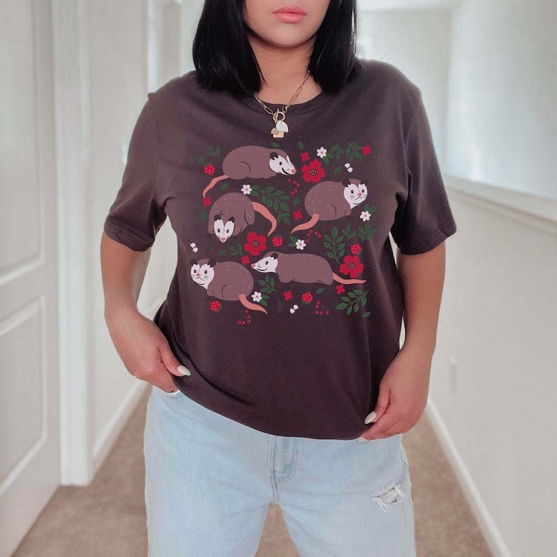 May include: A dark brown t-shirt with a graphic print of multiple opossums with red flowers and green leaves. The opossums are brown and white with black eyes.