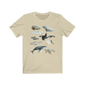 K&ouml;nnte beinhalten: Ein beiges T-Shirt mit einer Aquarell-Illustration verschiedener Meerestiere, darunter ein Wal, ein Delfin, eine Meeresschildkr&ouml;te, ein Seekuh, eine Robbe und ein Rochen.