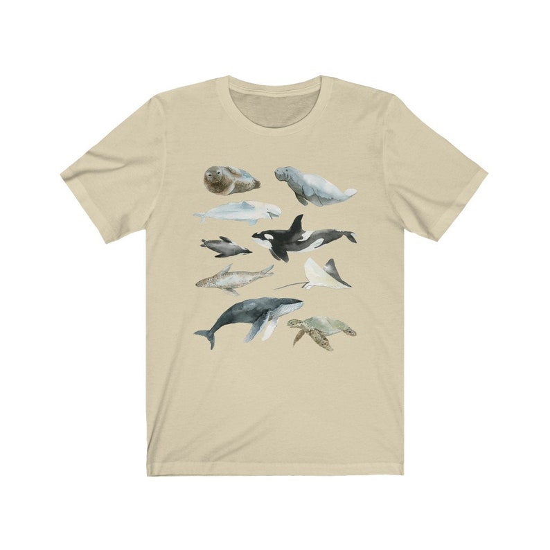 K&ouml;nnte beinhalten: Ein beiges T-Shirt mit einer Aquarell-Illustration verschiedener Meerestiere, darunter ein Wal, ein Delfin, eine Meeresschildkr&ouml;te, ein Seekuh, eine Robbe und ein Rochen.
