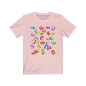 May include: A pink t-shirt with a colorful macaron pattern. The macarons are in various shades of pink, blue, green, yellow, and orange.