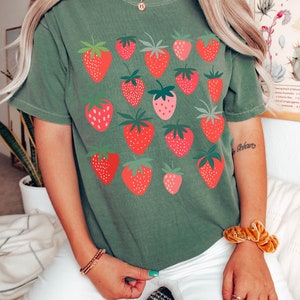 Puede incluir: Una camiseta verde con un estampado gr&aacute;fico de fresas rojas. La camiseta tiene mangas cortas y cuello redondo.
