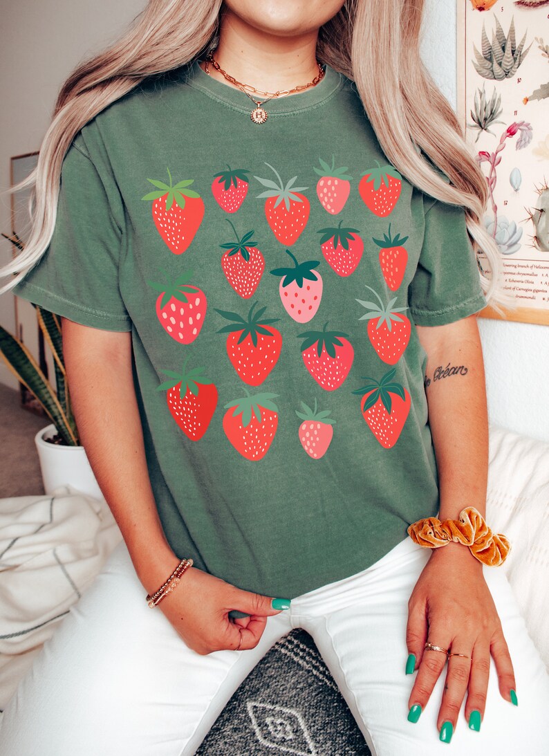 Puede incluir: Una camiseta verde con un estampado gr&aacute;fico de fresas rojas. La camiseta tiene mangas cortas y cuello redondo.