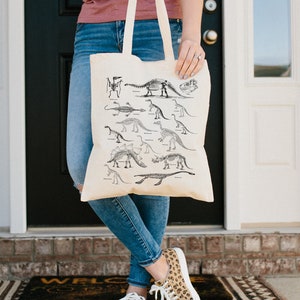 May include: A beige tote bag with a black and white illustration of dinosaur skeletons. The bag has two handles and is being held by a person wearing blue jeans.